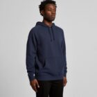 Man wearing a navy blue hoodie and black pants, standing against a plain gray background.