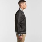 Man in a black bomber jacket and khaki pants standing in profile against a plain background.