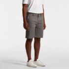 Man wearing a white shirt, gray shorts, and white sneakers standing against a plain background.