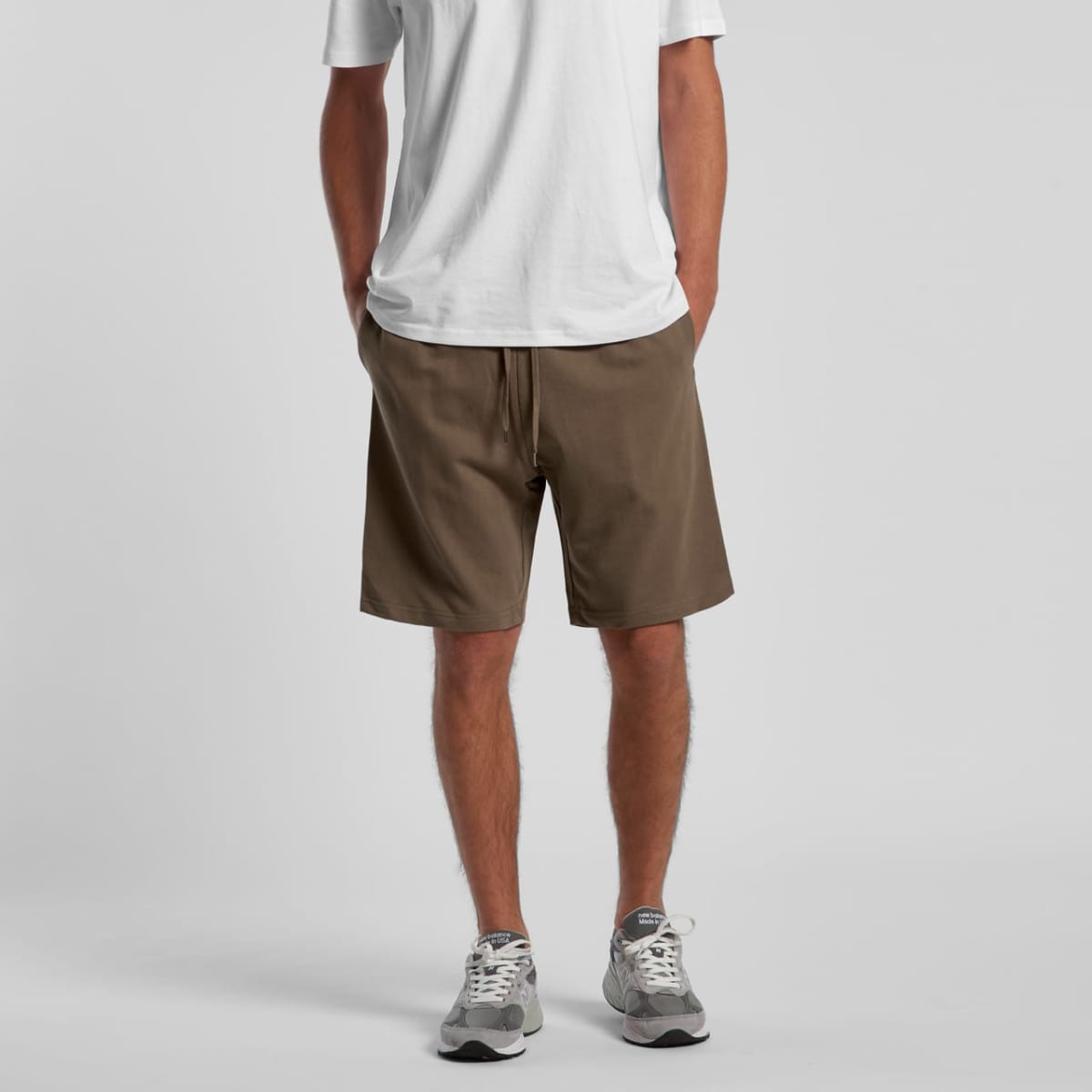 Man wearing a white t-shirt, brown shorts, and gray sneakers, standing with hands in pockets.