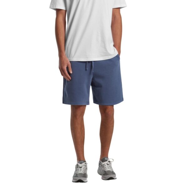 Man in white t-shirt, blue shorts, and gray sneakers standing with hands in pockets.