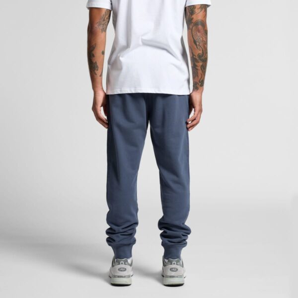 Man in white shirt and blue joggers standing, shown from the back, on a plain background.