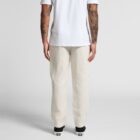 Man in white t-shirt and cream pants, seen from behind, shows tattooed arms and black shoes.