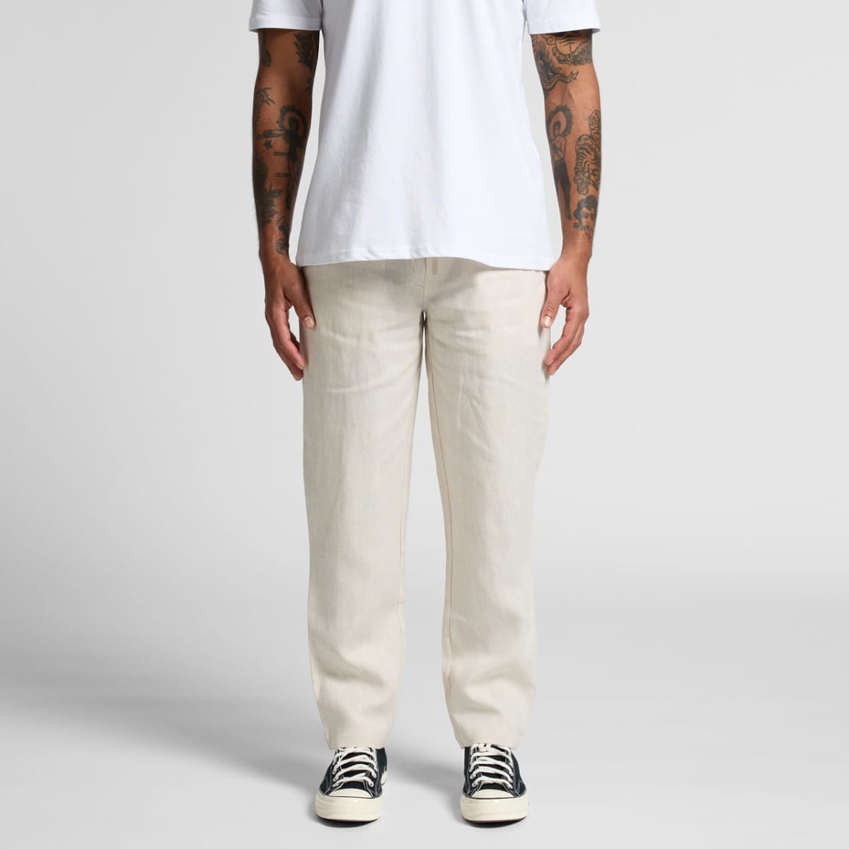 Person in white t-shirt, cream pants, and black sneakers standing against plain background.