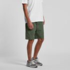 A person wearing a white shirt, green shorts, and gray sneakers stands against a plain backdrop.