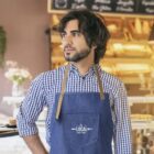 Man in blue checkered shirt and denim apron standing in a bakery, looking to the side.