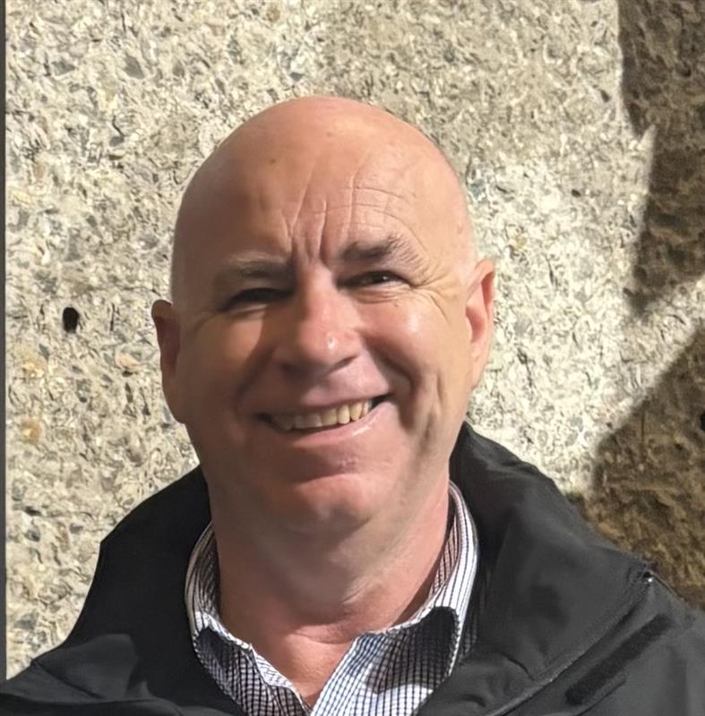 A smiling bald man in a black jacket stands before a stone wall—Wollongong Businesses welcome.