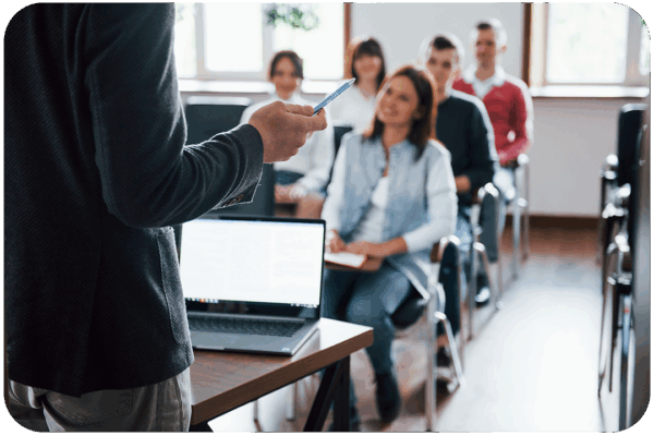 A person presents at a small conference to a group seated in a classroom with a laptop.