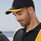 Man in black and yellow cap looking down at a camera, outdoors with blue sky in background.