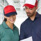 Two people wearing caps look at a sheet of paper together in a bright hallway.