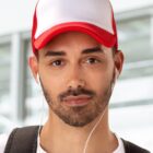 Man with a red and white cap, wearing earphones and a backpack, looking at the camera.