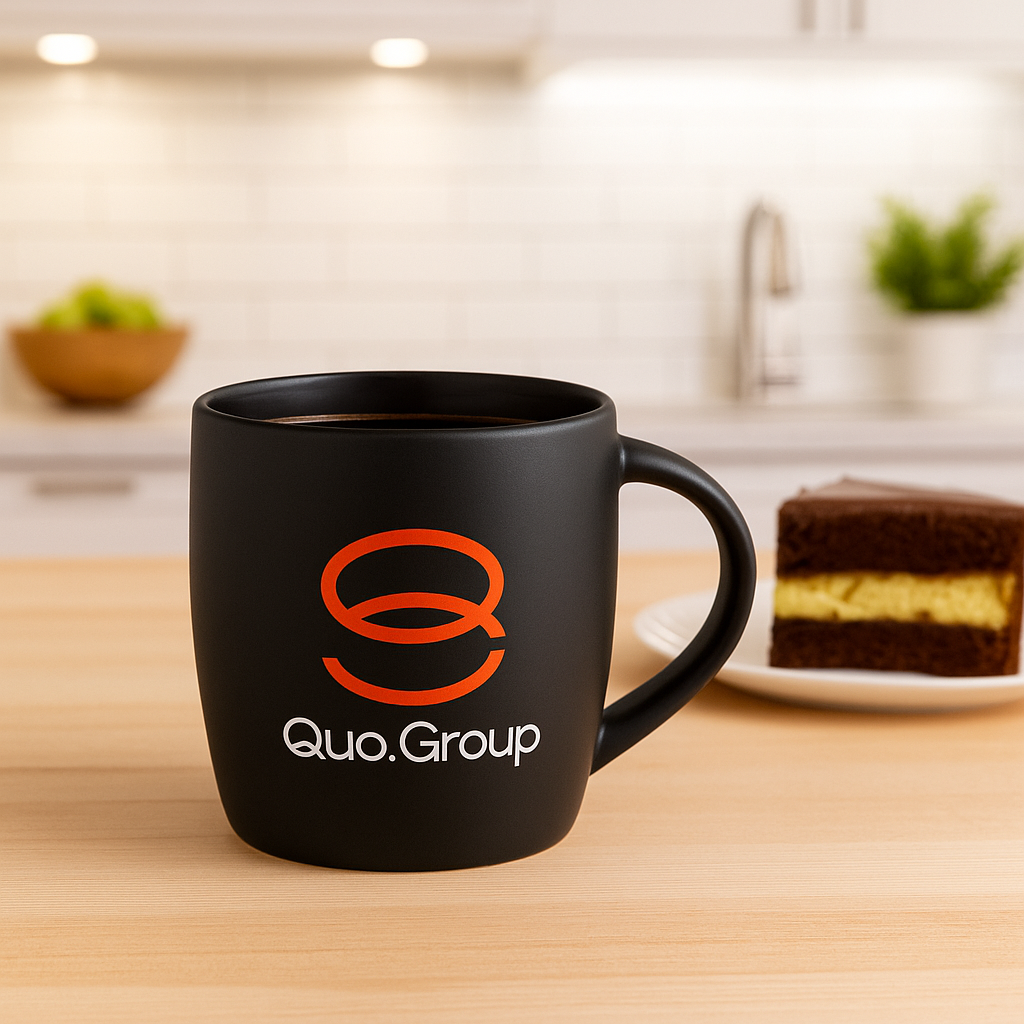 Black mug with Quo.Group logo—simple branded gift idea, cake slice on plate behind.