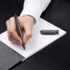 A hand writing on a notepad with a pen; pen cap rests nearby on the black desk.