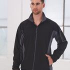 Man wearing a black zip-up jacket stands indoors in front of large windows.