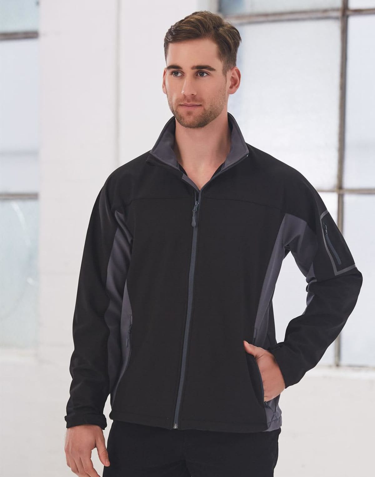 Man wearing a black zip-up jacket stands indoors in front of large windows.
