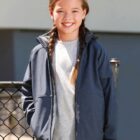 Girl with braided hair in a gray jacket, smiling and standing by a chain-link fence.
