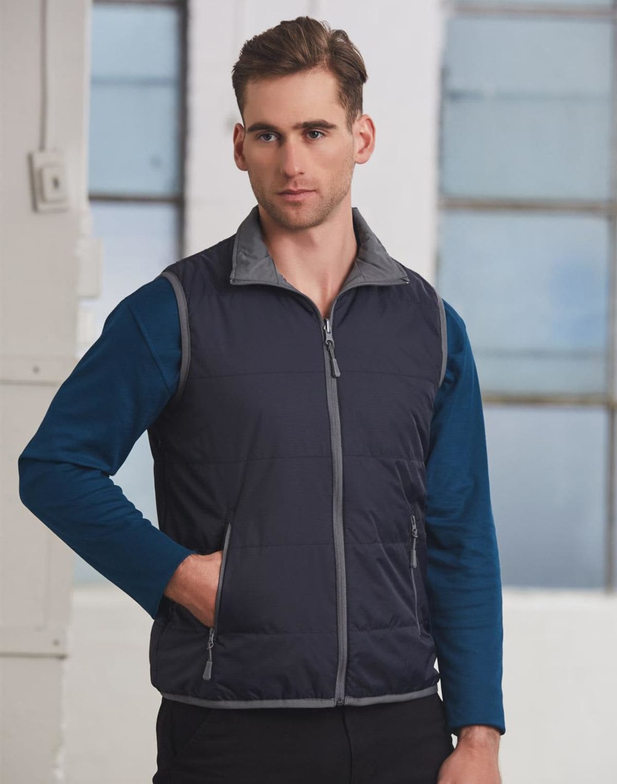 Man wearing a navy blue zip-up vest over a blue long-sleeve shirt stands indoors.