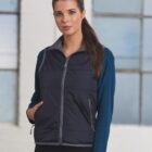 Woman wearing a navy vest over a blue long-sleeve shirt stands in front of large windows.