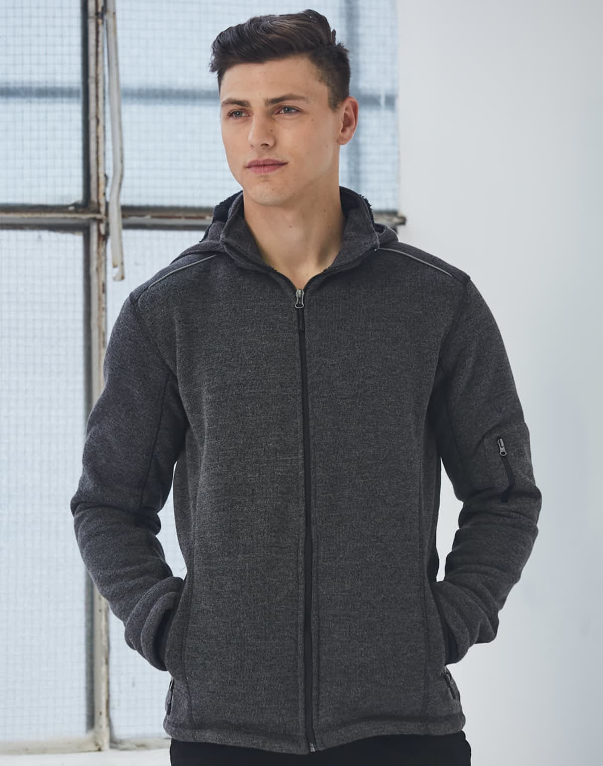 Man in a dark gray zip-up hoodie standing indoors with hands in pockets.