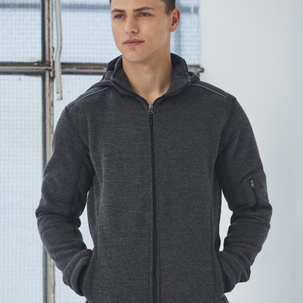 Man in a dark gray zip-up jacket standing indoors with hands in pockets.