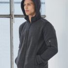Man in a dark gray zip-up hoodie stands indoors by a window, looking to the side.