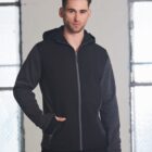 Man wearing a black and gray hooded zip-up jacket standing indoors by large windows.