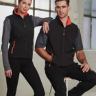 Two people wearing matching black vests with orange accents, posing indoors by a window.
