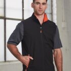 Man wearing a black vest with orange accents over a gray shirt, standing indoors by windows.
