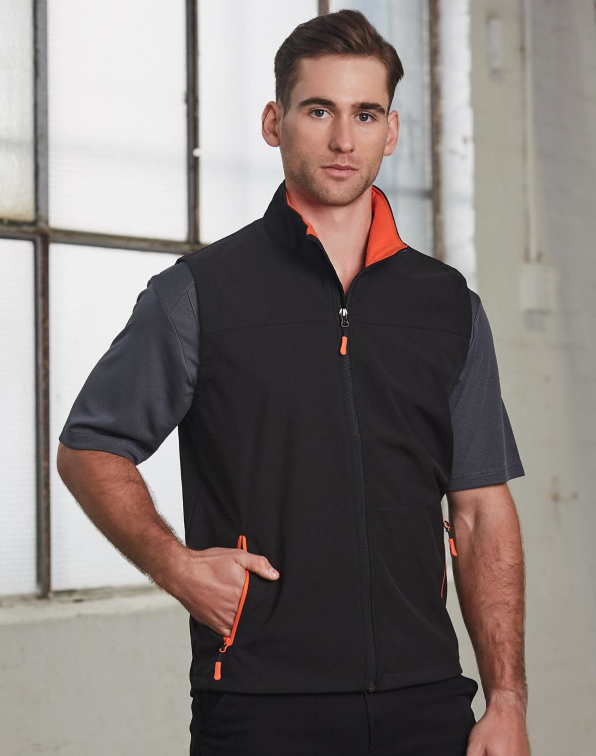 Man wearing a black vest with orange accents over a gray shirt, standing indoors by windows.