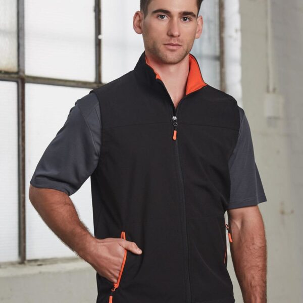 Man wearing a black and gray zip-up vest with orange accents, standing indoors by a window.