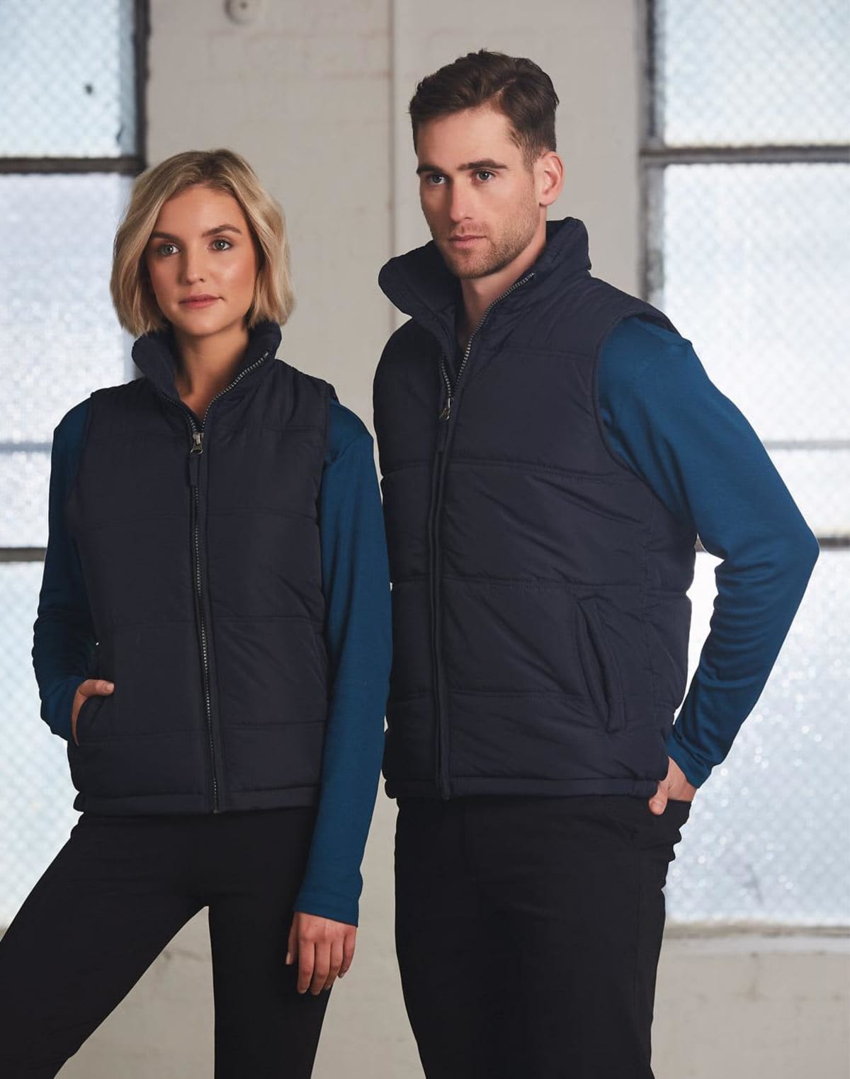 Two people wear navy vests over blue shirts, standing indoors in front of large windows.
