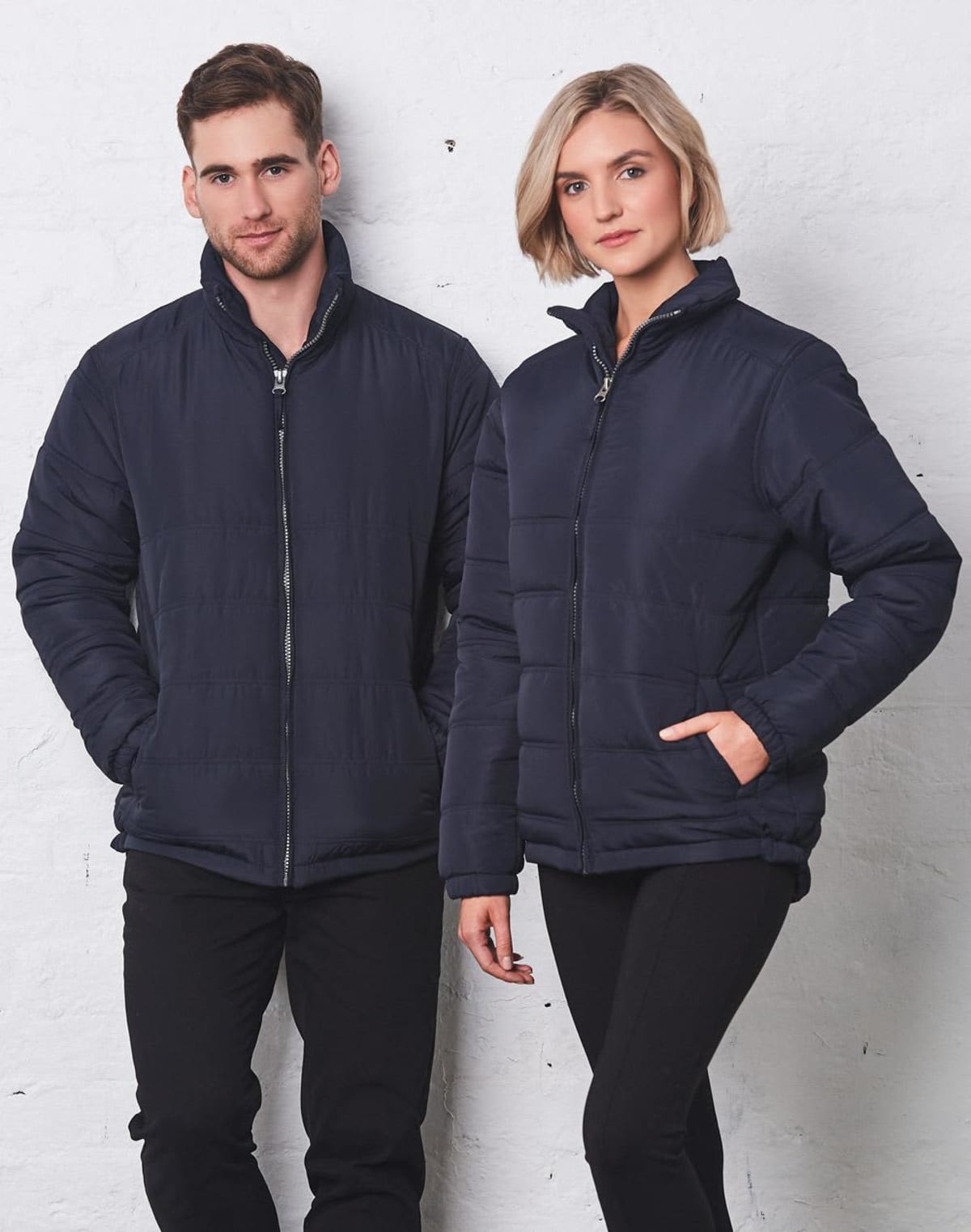 Man and woman in navy puffer jackets stand against a white wall, posing together.