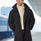 Man in a black coat and cream scarf standing on a train platform.