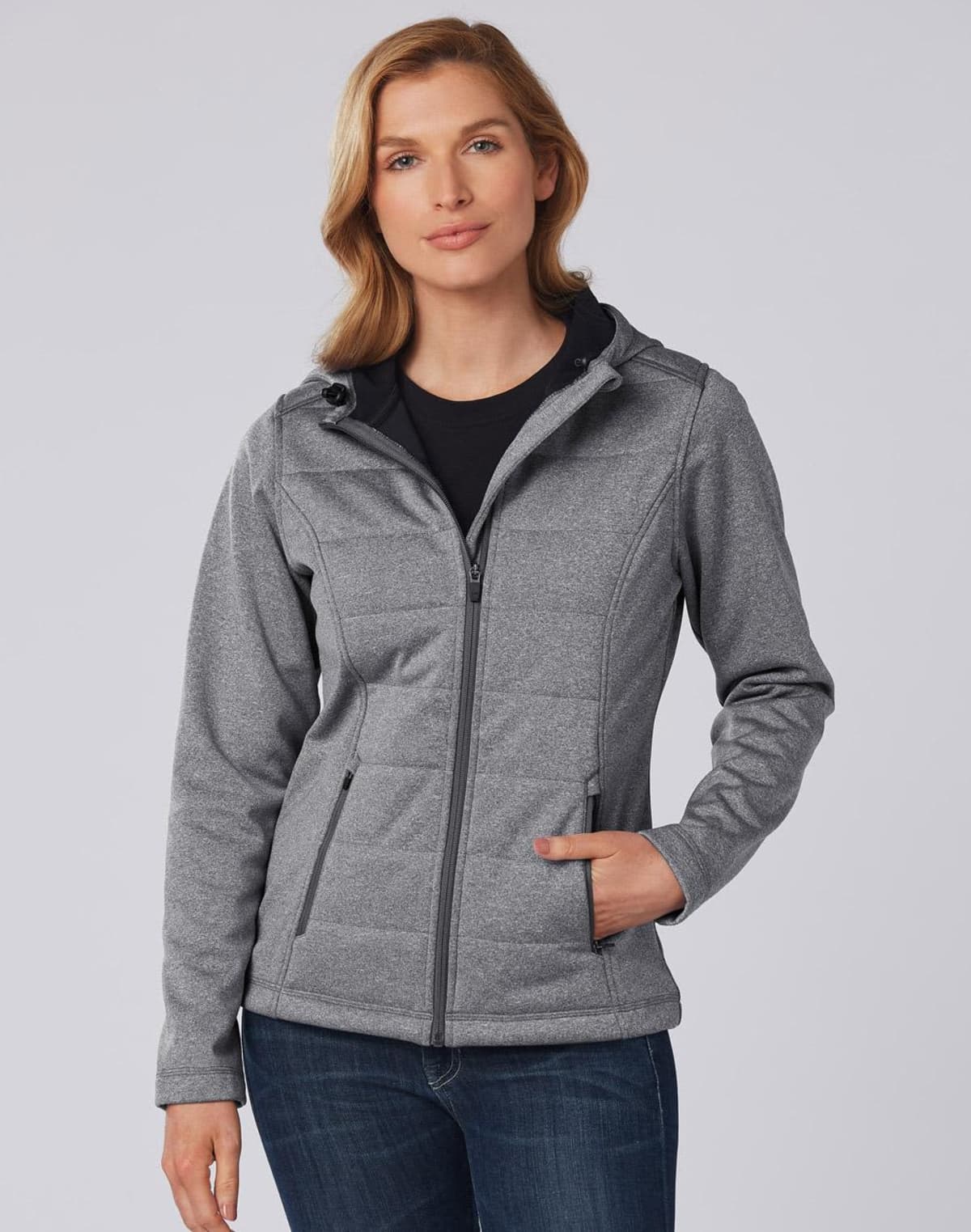 Woman wearing a gray zip-up jacket with a hood and blue jeans, standing against a plain background.