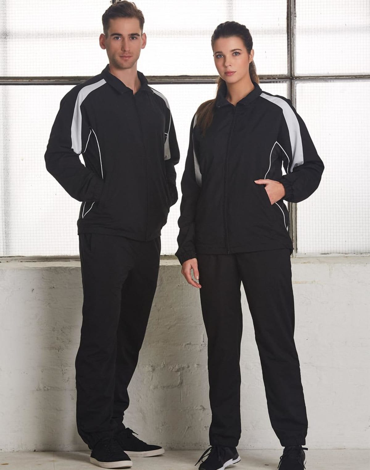 Two people wearing matching black and white tracksuits standing indoors by a window.