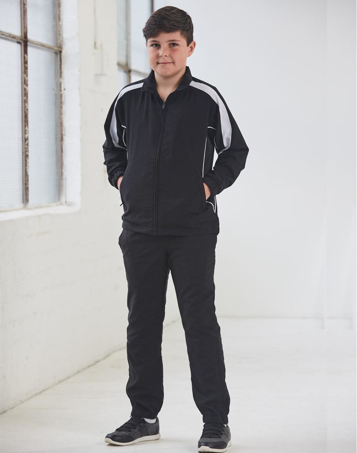 A boy in a black tracksuit stands with hands in pockets in a bright, white room.