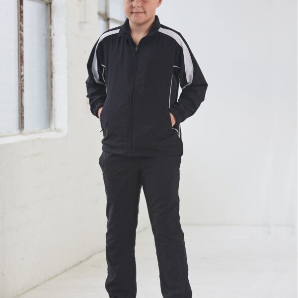 A boy in a black tracksuit stands indoors with his hands in his pockets, smiling slightly.