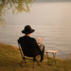 Person in a black hat reads a book by the water, sitting on a chair near a small table.