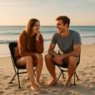 Two people sit on chairs laughing together on a sandy beach at sunset.