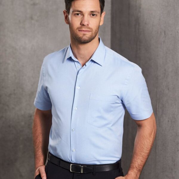Man in a light blue short-sleeve shirt and black pants standing against a gray background.
