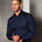 Man in a dark blue dress shirt standing against a textured gray wall.