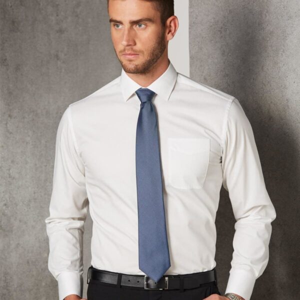 Man in a white dress shirt, blue tie, and black pants standing against a gray wall.