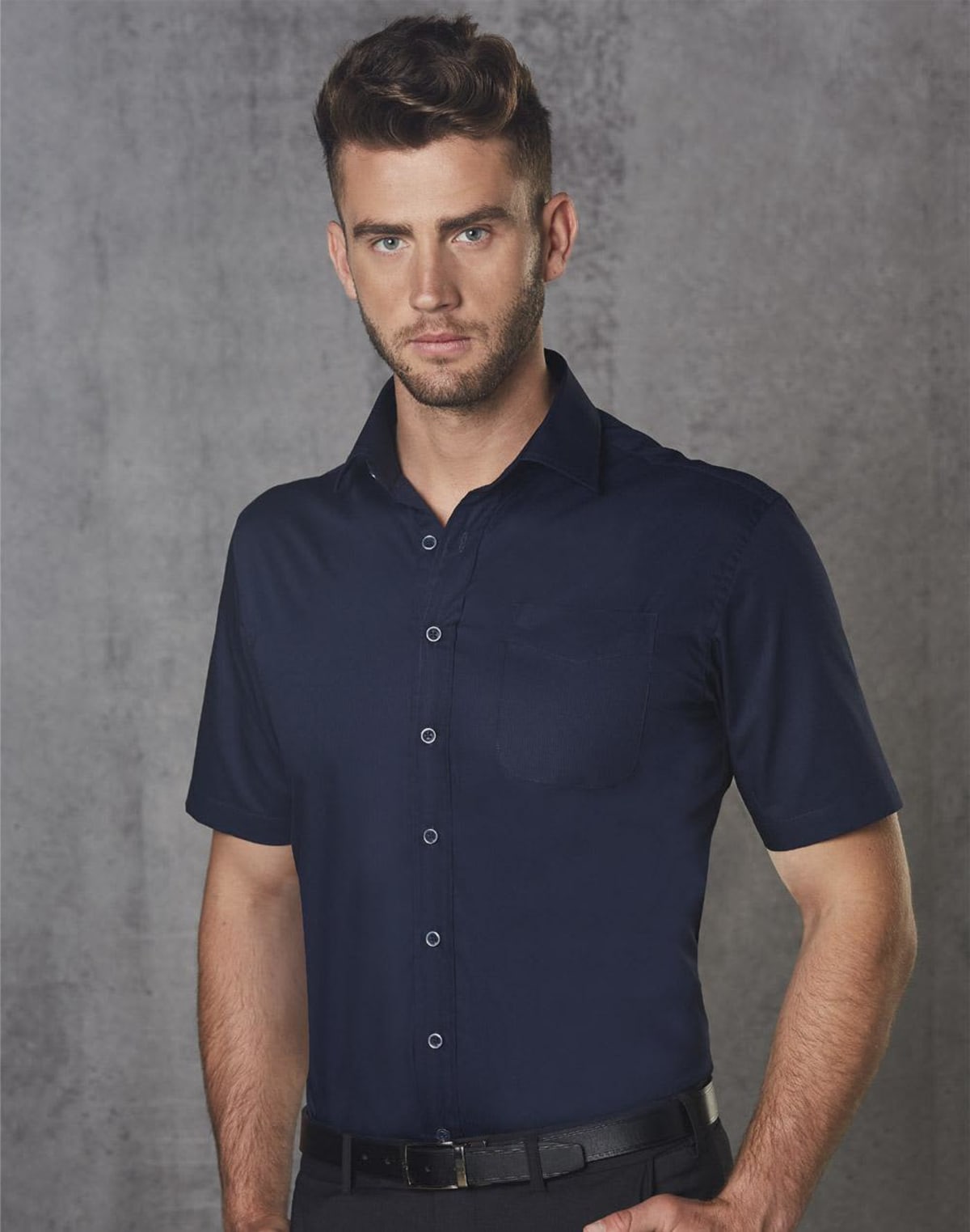 Man in a dark blue short-sleeved shirt standing against a gray textured background.