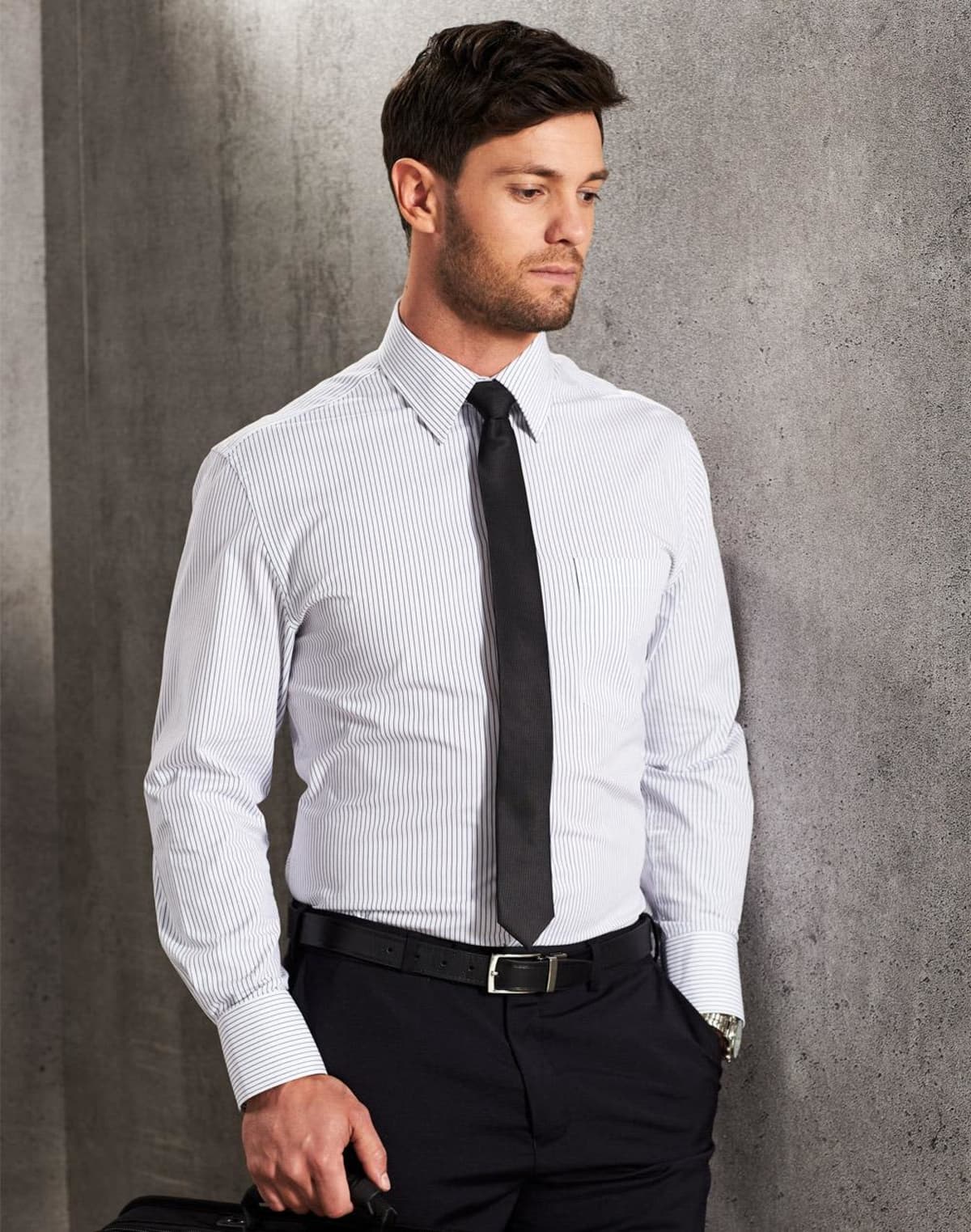 Man in striped dress shirt, black tie, and slacks standing against a gray concrete wall.