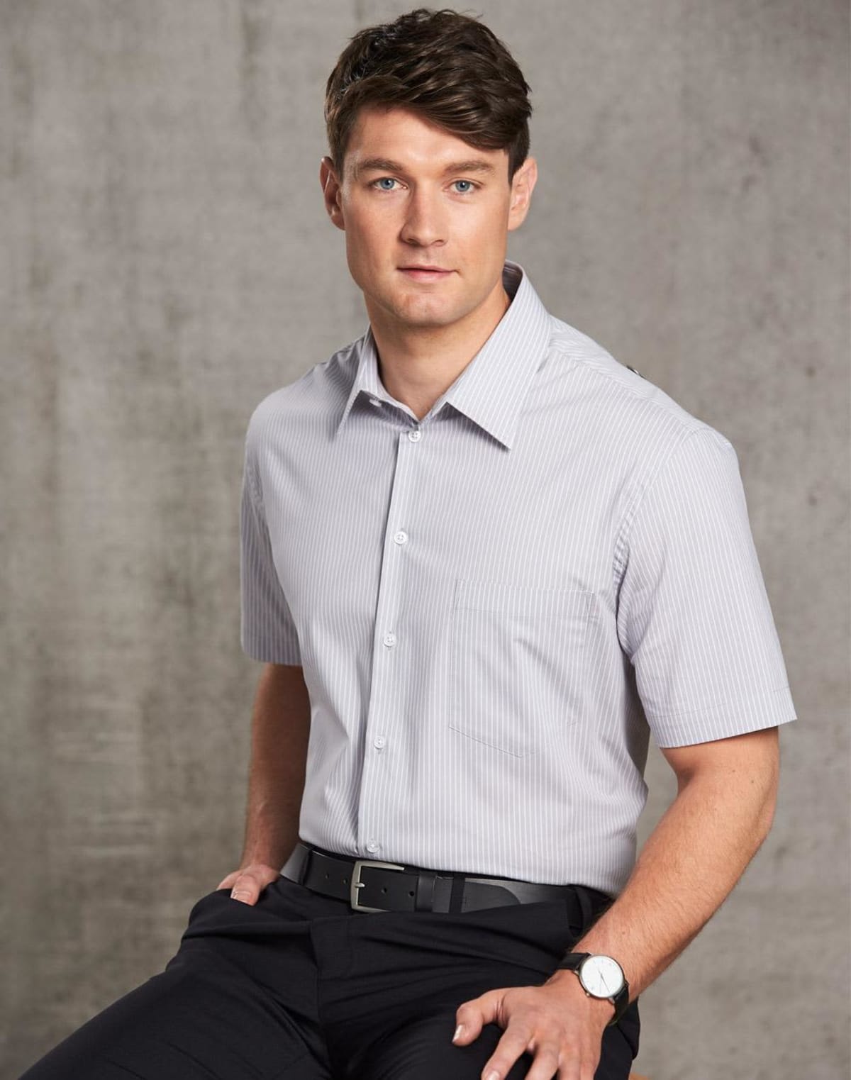 Man in a light gray short-sleeve dress shirt and black pants sitting against a gray background.