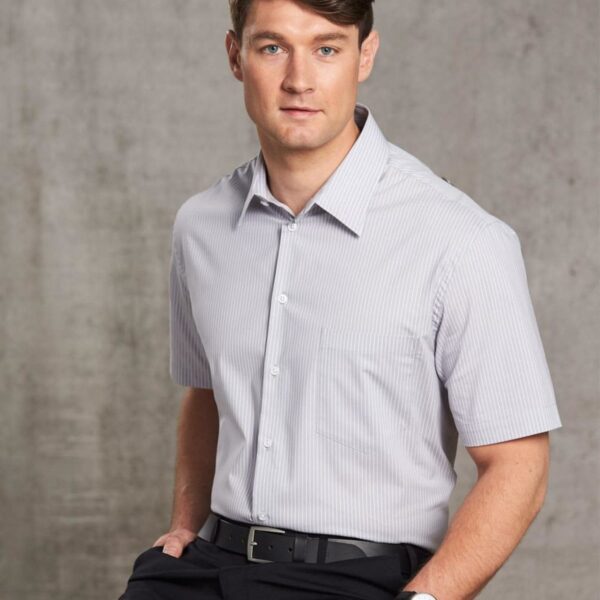 Man in a short-sleeve dress shirt and black pants sitting against a gray textured background.