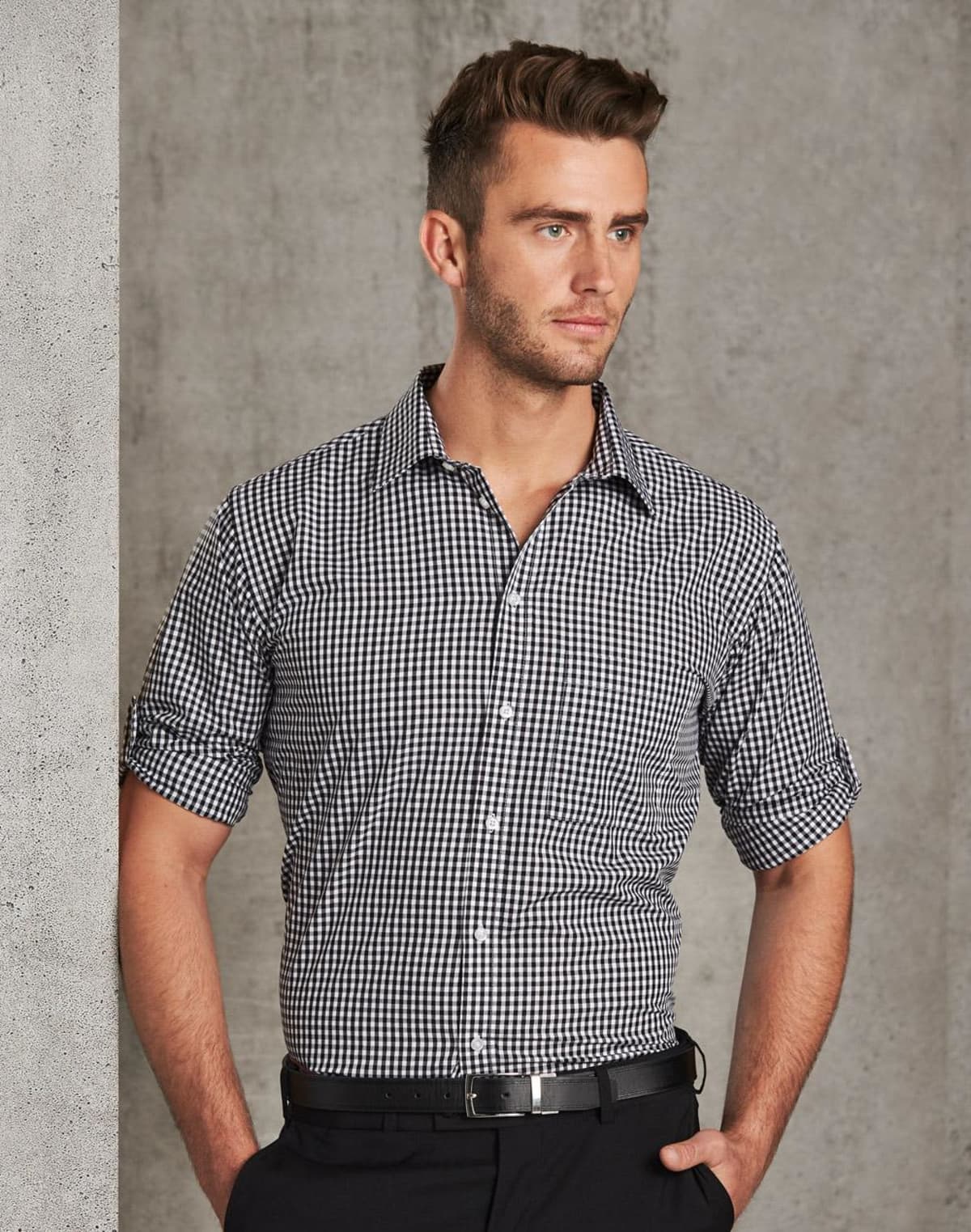 Man in a checkered shirt standing against a gray textured wall, looking to the side.