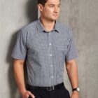 Man in a short-sleeved checkered shirt standing against a plain gray wall, looking right.