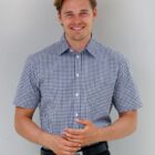 Smiling man in a short-sleeve checkered shirt standing against a plain light background.