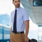 A man in a short-sleeve shirt and tie holds a bag at a train station platform.